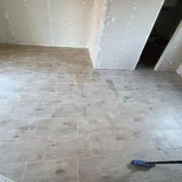 Ułożona podłoga z płytek imitujących drewno w pokoju, ściany przygotowane pod malowanie, widoczne ślady po szpachlowaniu, na podłodze leży niebieska szczotka.