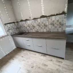 Montaż płytek ściennych z dekoracyjnym wzorem w kuchni, widoczne szare szafki pod blatem i płytki podłogowe imitujące drewno, ściany w trakcie wykończenia.