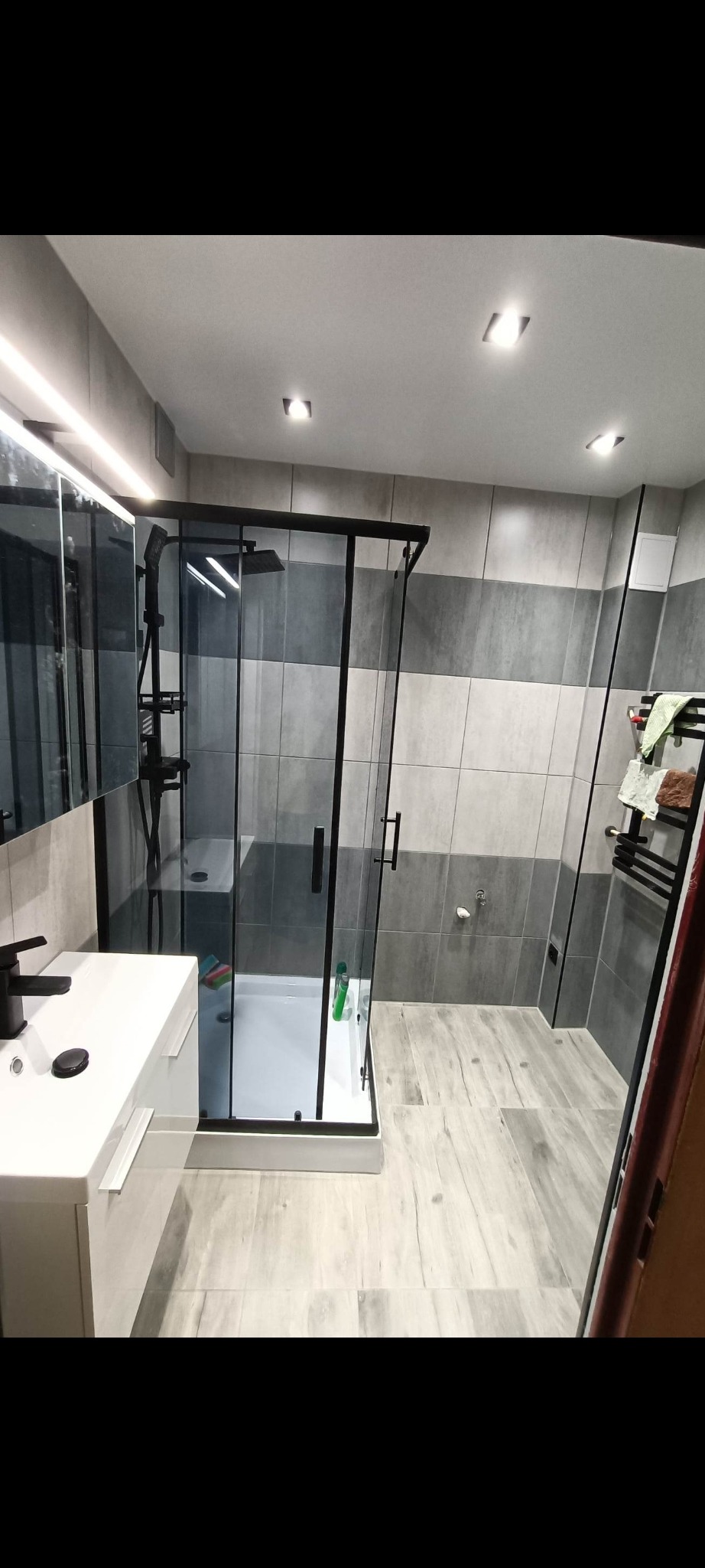 Nowoczesna łazienka z kabiną prysznicową typu walk-in, szare płytki na ścianach i podłodze imitujące drewno, biała umywalka z czarną armaturą, oświetlenie punktowe.