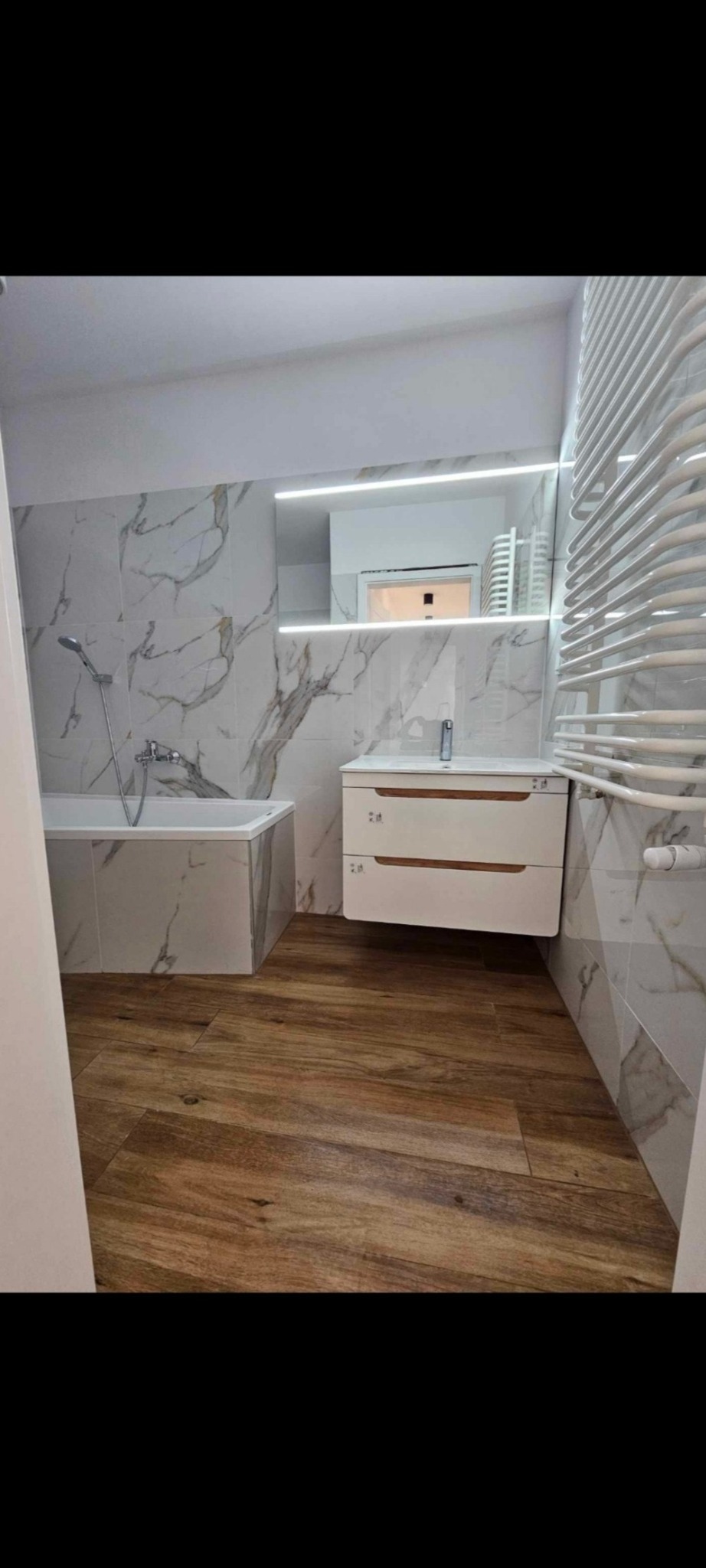 Nowoczesna łazienka po remoncie: drewnopodobna podłoga, białe marmurowe płytki, podświetlane lustro, biała szafka z drewnianymi akcentami, grzejnik drabinkowy.