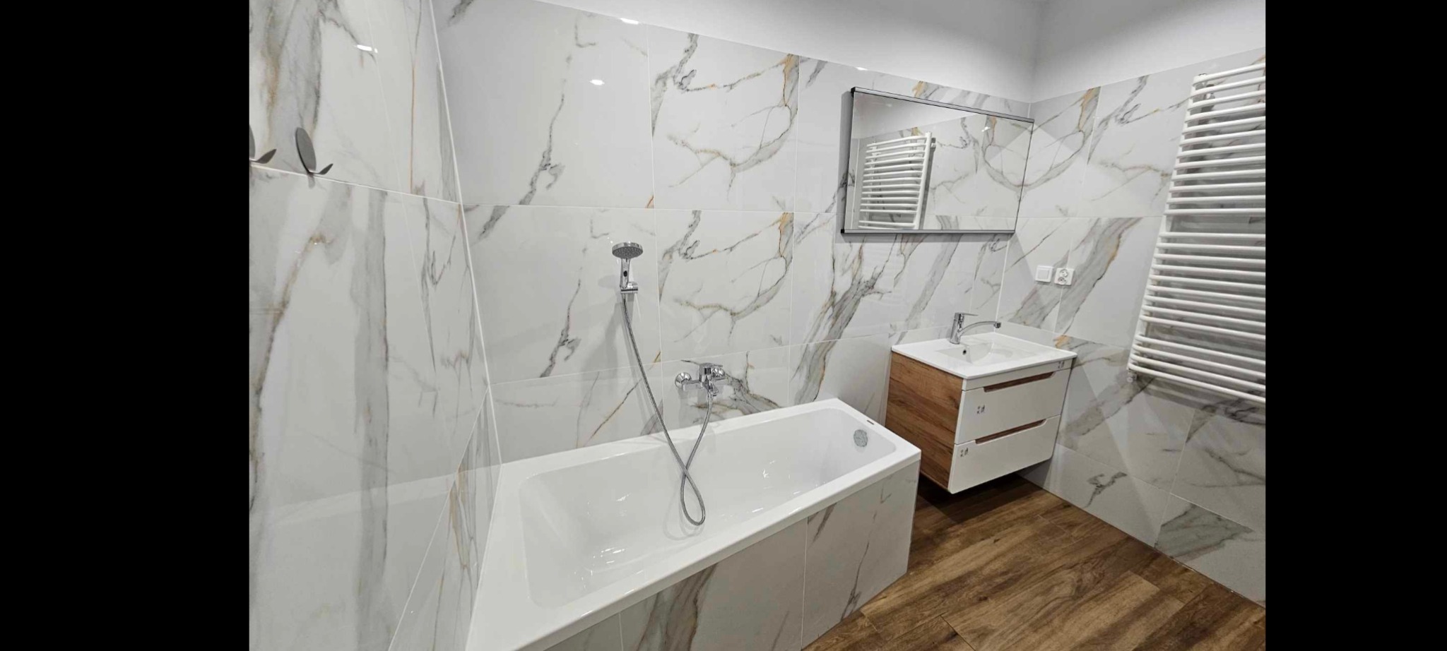 Biała łazienka z wanną, marmurowe płytki z brązowymi żyłami, drewnopodobna podłoga, szafka z umywalką i lustro z grzejnikiem.