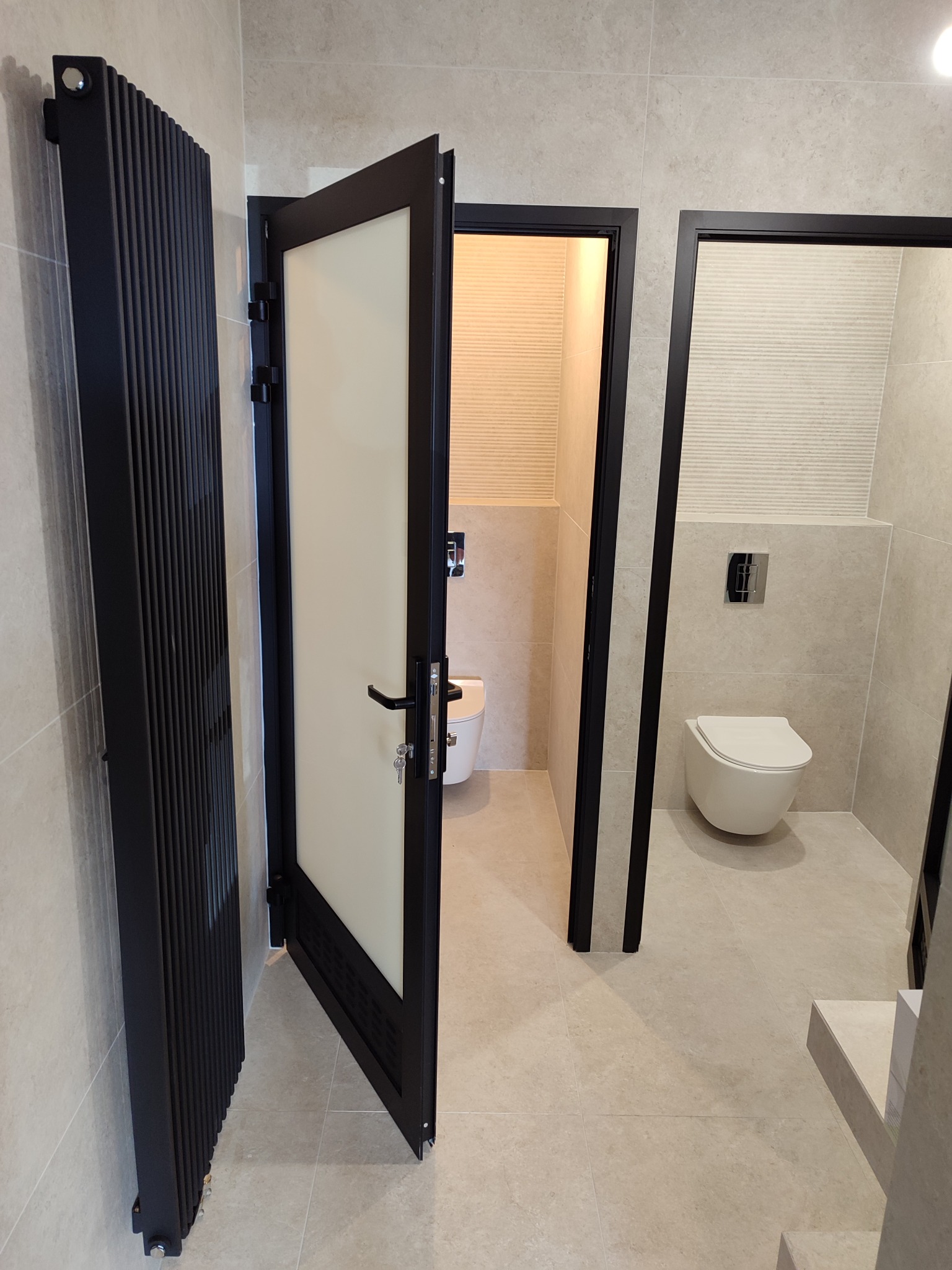 Nowoczesna łazienka z beżowymi płytkami, czarnym grzejnikiem żeberkowym i drzwiami z czarną ramą. Widok na toaletę w głębi przez otwarte drzwi.