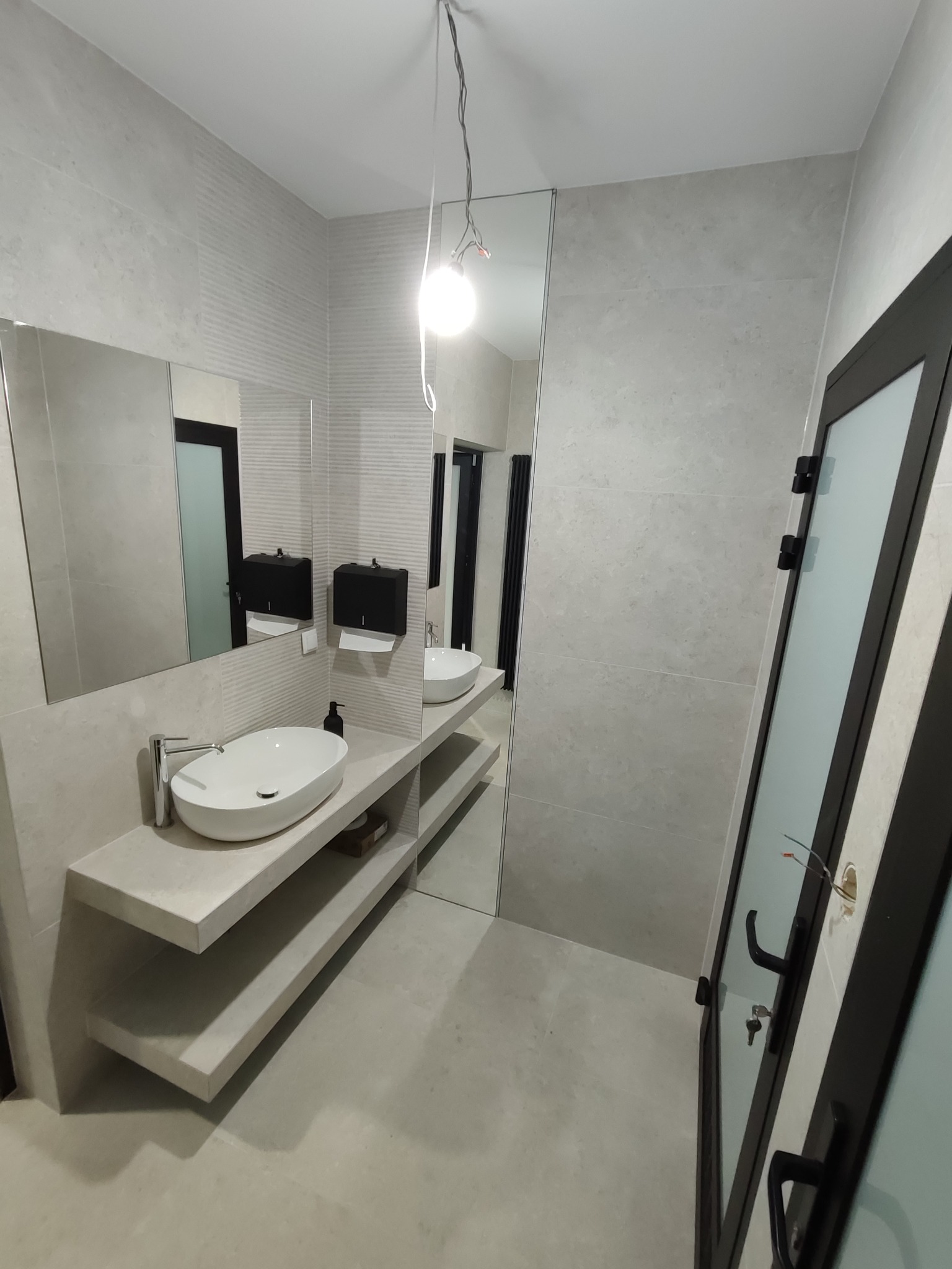 Nowoczesna łazienka z umywalką nablatową na półkach, duże lustro, szare płytki na ścianach i podłodze, czarne akcenty. Widoczna instalacja elektryczna przy drzwiach.