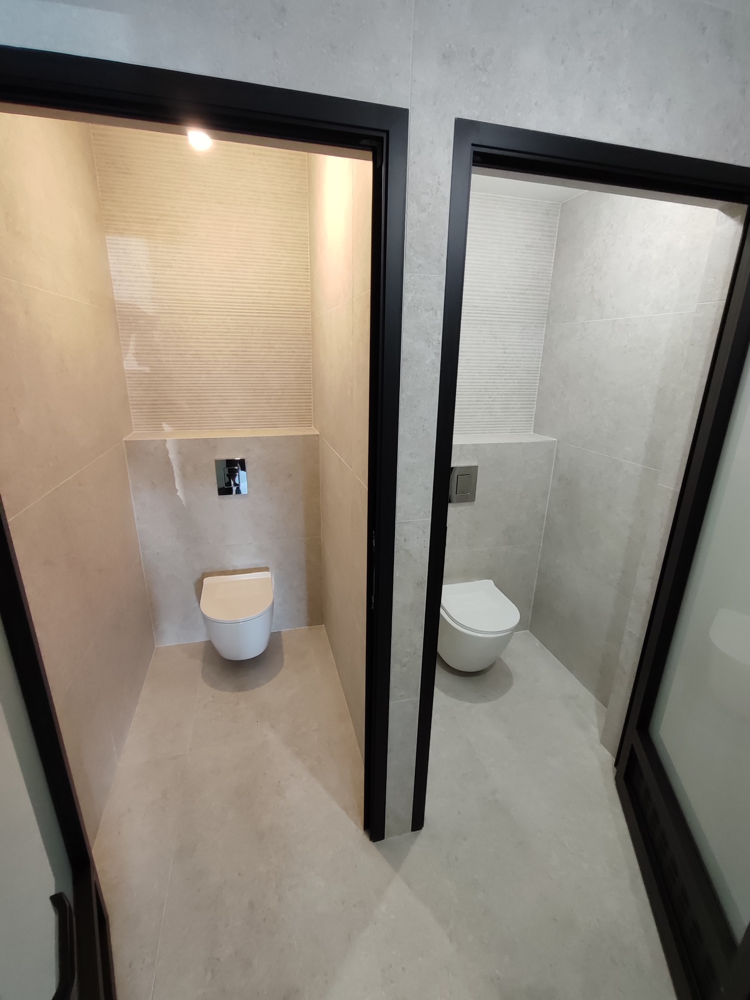 Dwa wykończone, nowoczesne WC z podwieszanymi miskami, jasnymi płytkami i czarnymi ramami drzwi, widoczne z perspektywy korytarza. Minimalistyczny design.