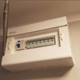 Biała skrzynka rozdzielcza z otwartą klapą, zamontowana na ścianie, widoczne zabezpieczenia i wyłączniki, lekko niedoświetlone zdjęcie.