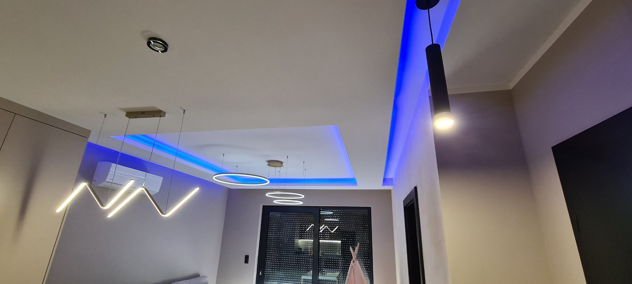 Nowoczesne oświetlenie sufitowe z niebieskim podświetleniem LED i lampami wiszącymi w salonie w Zielonej Górze. Widoczny klimatyzator i minimalistyczny wystrój.