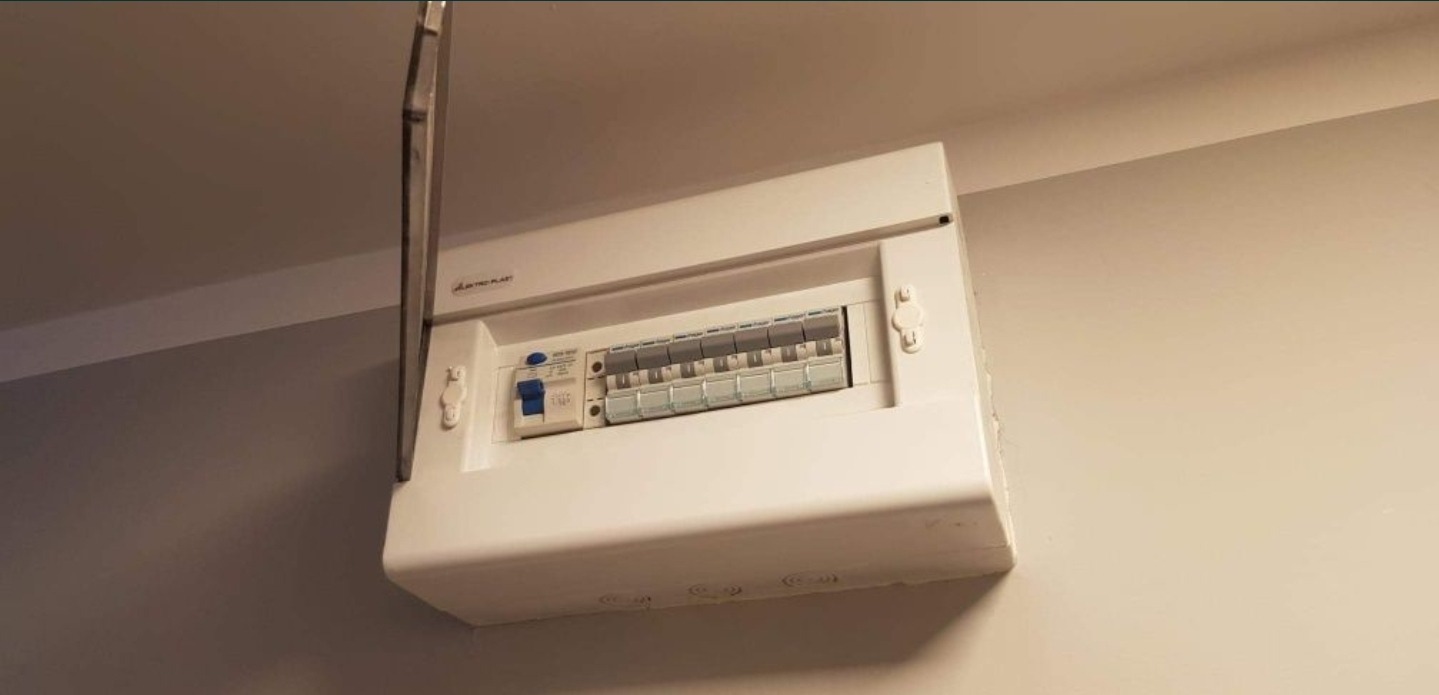 Biała skrzynka rozdzielcza z otwartą klapą, zamontowana na ścianie, widoczne zabezpieczenia i wyłączniki, lekko niedoświetlone zdjęcie.