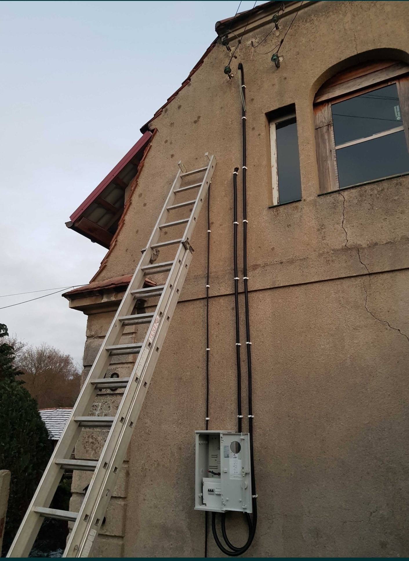 Zewnętrzna instalacja elektryczna na ścianie budynku z drabiny, widoczna skrzynka rozdzielcza z otwartymi drzwiczkami i kable w peszlach przymocowane do elewacji.