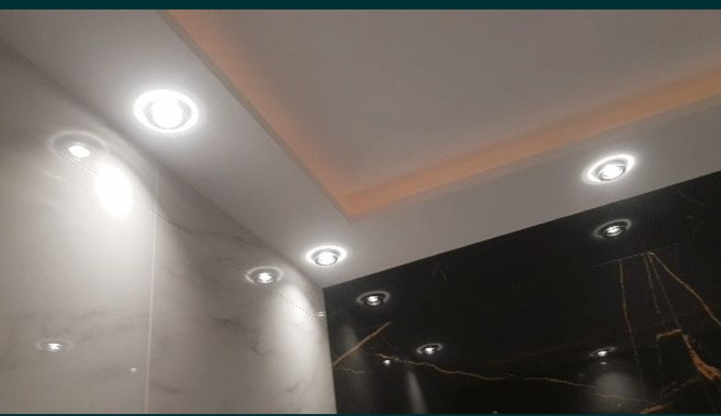 Nowoczesna łazienka z marmurowymi ścianami w odcieniach bieli i czerni, oświetlona punktowymi lampami sufitowymi i subtelnym oświetleniem LED na suficie podwieszanym.