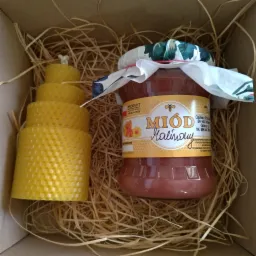 Słoik z miodem malinowym i świeca z wosku pszczelego ułożone w pudełku wypełnionym siankiem.