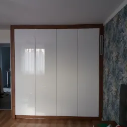 Biała szafa wnękowa z drewnianą ramą, widoczny fragment pokoju z wzorzystą tapetą i osobą siedzącą w oddali.