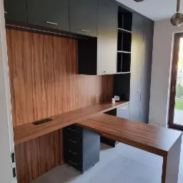 Nowoczesne biuro domowe z drewnianym blatem w kształcie litery L, czarnymi szafkami górnymi i szufladami, oraz szarą podłogą z płytek, oświetlone wiszącymi lampami z przydymionego szkła.