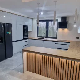 Nowoczesna kuchnia z szarymi frontami szafek, drewnianym blatem i panelem dekoracyjnym z pionowych listew, czarna lodówka side-by-side i oświetlenie punktowe.