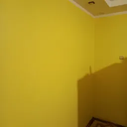Żółta ściana z dekoracyjną gładzią w rogu pokoju, widoczny fragment sufitu z oświetleniem i fragment podłogi z wzorzystymi płytkami.