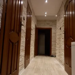 Wnętrze korytarza z trzema drewnianymi drzwiami o dekoracyjnych elementach, wykończonego płytkami imitującymi kamień na ścianach i podłodze, oświetlonego punktowymi lampami sufitowymi.
