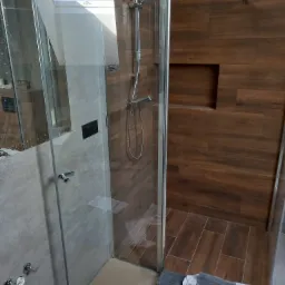 Nowoczesna kabina prysznicowa z szarymi płytkami imitującymi kamień i panelami ściennymi imitującymi drewno, widoczne niedokończone prace wykończeniowe przy podłodze.