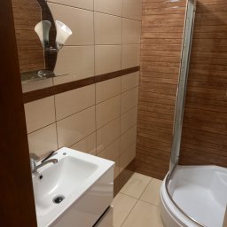 Technika Budowlana Mariusz Ogorzałek - Umywalka z chromowaną baterią i lustrem z półką w łazience wyłożonej beżowymi i brązowymi płytkami imitującymi drewno oraz kabina prysznicowa.