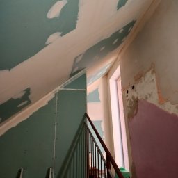 Technika Budowlana Mariusz Ogorzałek - Poddasze w trakcie wykańczania: widoczne płyty gipsowo-kartonowe na skośnym suficie i ścianach, częściowo pokryte masą szpachlową, drabina aluminiowa, fragment schodów z metalową barierką, surowe...