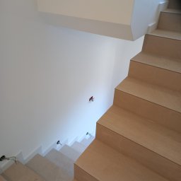 Technika Budowlana Mariusz Ogorzałek - Wykończone schody wewnętrzne z okładziną z płytek imitujących drewno, widoczne punkty oświetleniowe w ścianie i na schodach. Minimalistyczny design wnętrza.