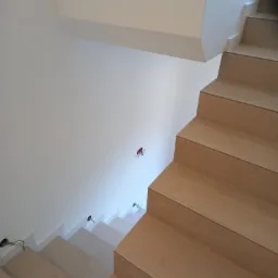 Wykończone schody wewnętrzne z okładziną z płytek imitujących drewno, widoczne punkty oświetleniowe w ścianie i na schodach. Minimalistyczny design wnętrza.