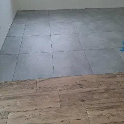 Połączenie szarych płytek podłogowych i paneli imitujących drewno na podłodze w trakcie prac wykończeniowych, widoczne otwory instalacyjne w ścianie.