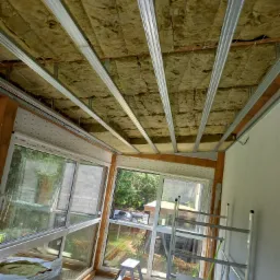 Ocieplenie poddasza wełną mineralną z widoczną konstrukcją metalową. W tle okna z widokiem na zieleń. Na pierwszym planie drabina i materiały izolacyjne Rockwool.