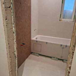 Technika Budowlana Mariusz Ogorzałek - Wnętrze łazienki w trakcie prac wykończeniowych: częściowo ułożone płytki imitujące drewno, wanna obudowana płytkami, okno. Widoczne elementy instalacji i niezakończone obramowanie drzwi.