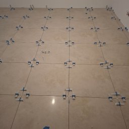 Technika Budowlana Mariusz Ogorzałek - Ułożone beżowe płytki podłogowe z systemem poziomowania, widoczne fugi i elementy systemu. Fragment ściany i wystające przewody elektryczne w tle.