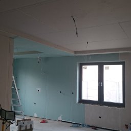 Technika Budowlana Mariusz Ogorzałek - Wnętrze w trakcie montażu płyt karton-gips na ścianach i suficie. Widoczne przewody elektryczne, drabina i narzędzia. Okno z czarną ramą. Surowy stan wykończenia.
