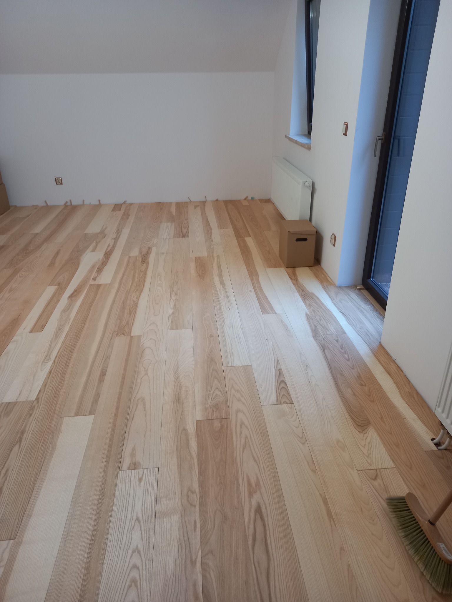 Jasna, drewniana podłoga ułożona w pokoju z białymi ścianami, widoczne listwy dystansowe przy ścianach, kartonowe pudełko i fragment grzejnika przy oknie.