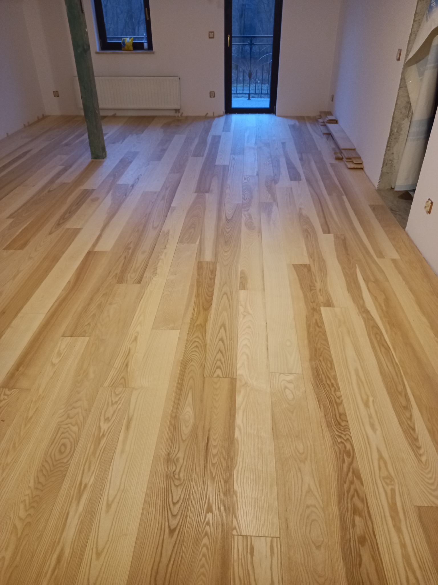 Nowo położona, jasna, drewniana podłoga w pokoju z widocznym słupem konstrukcyjnym i wyjściem na balkon.