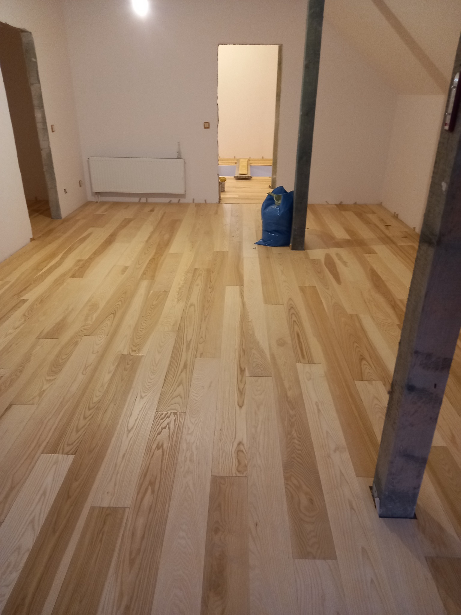Jasna, drewniana podłoga ułożona w pokoju na poddaszu z widocznymi drewnianymi słupami konstrukcyjnymi i niebieskim workiem z materiałami budowlanymi w tle.