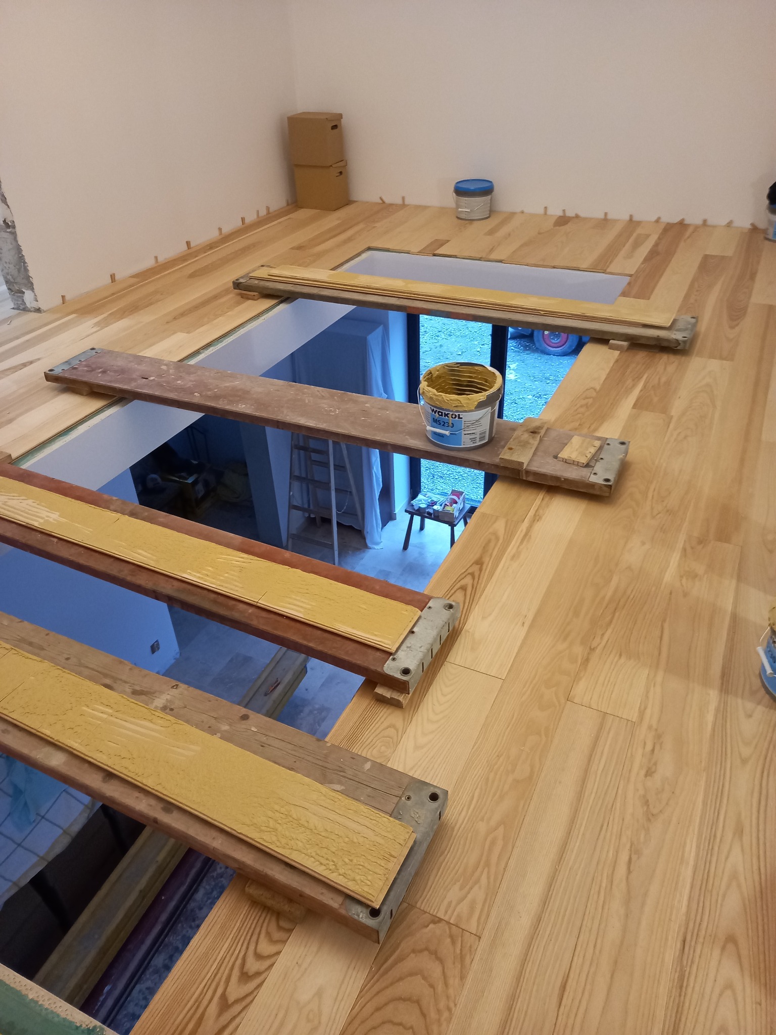 Układanie jasnej drewnianej podłogi z przerwą na schody, widoczne deski podtrzymujące i wiadro kleju Wakol MS 230.