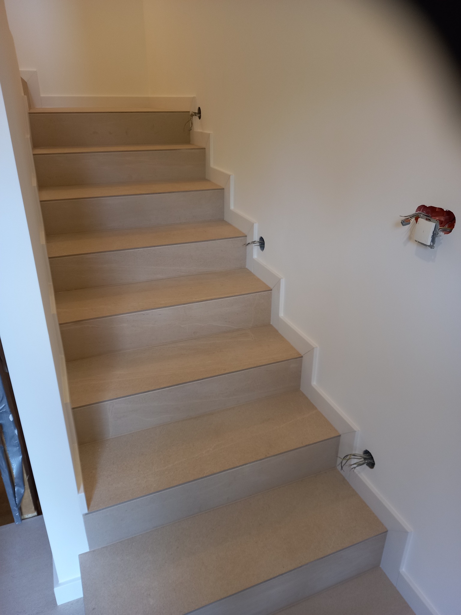 Wykończone schody z jasnymi stopniami, białe listwy przypodłogowe, widoczne przygotowania do montażu oświetlenia na ścianie.