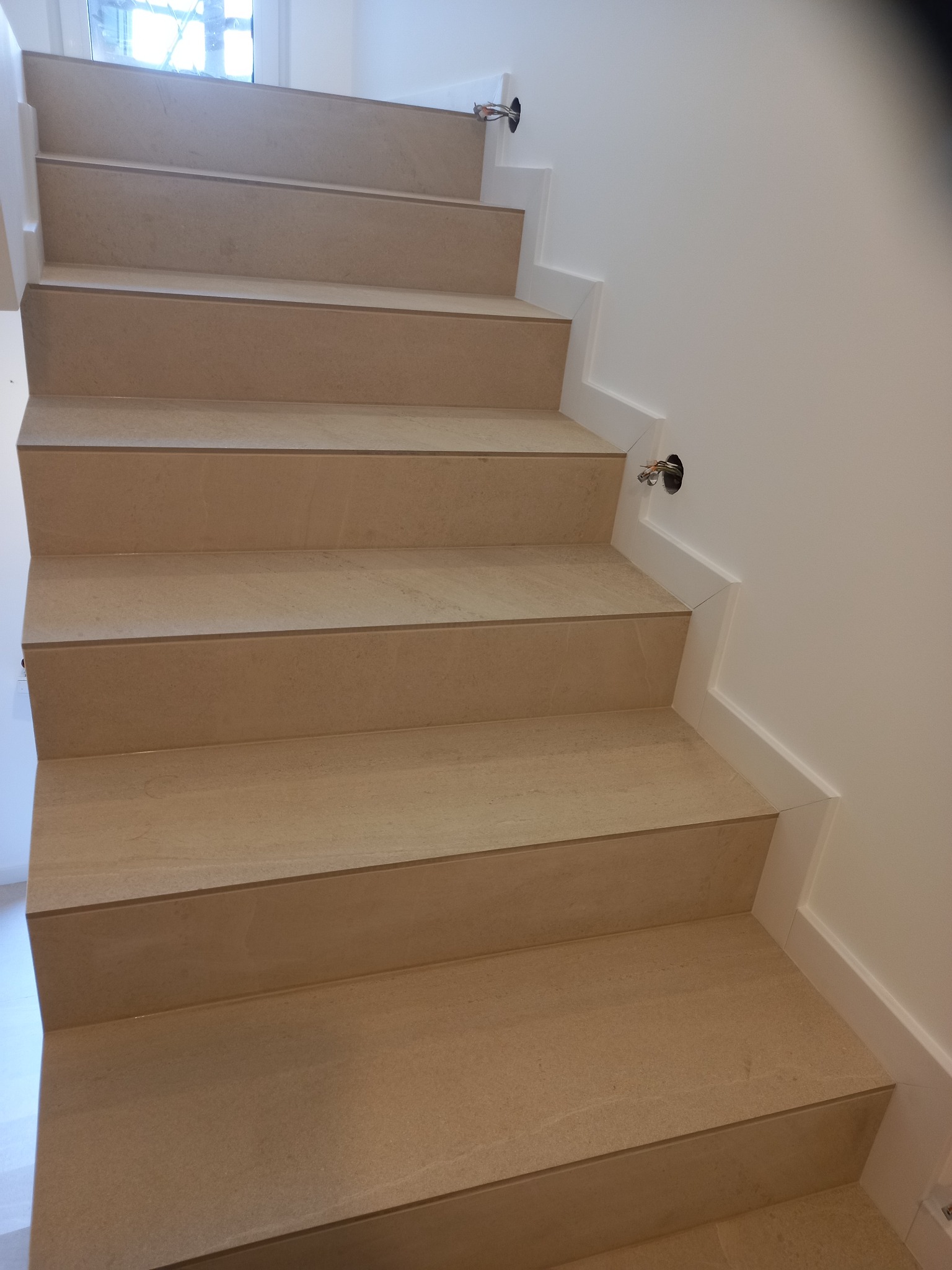 Wykończone schody z beżowych płytek, widoczne otwory na oświetlenie w ścianie. Minimalistyczne białe wykończenie krawędzi. Ujęcie z dołu, perspektywa w górę.