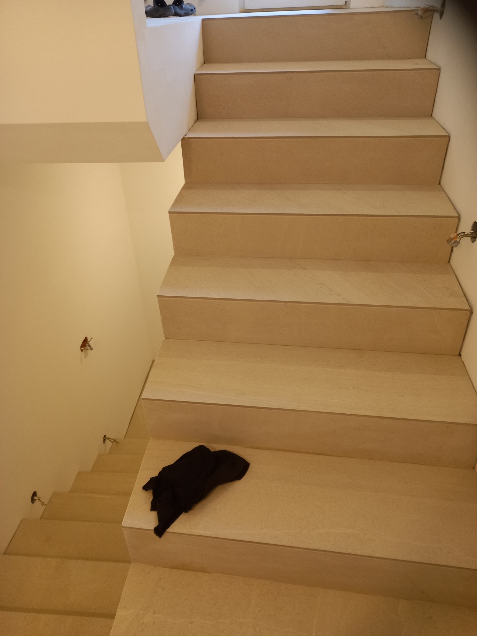 Wykończone schody z jasnego kamienia, widok z góry, z czarnym materiałem leżącym na jednym ze stopni, oraz fragmentem instalacji elektrycznej na ścianie.