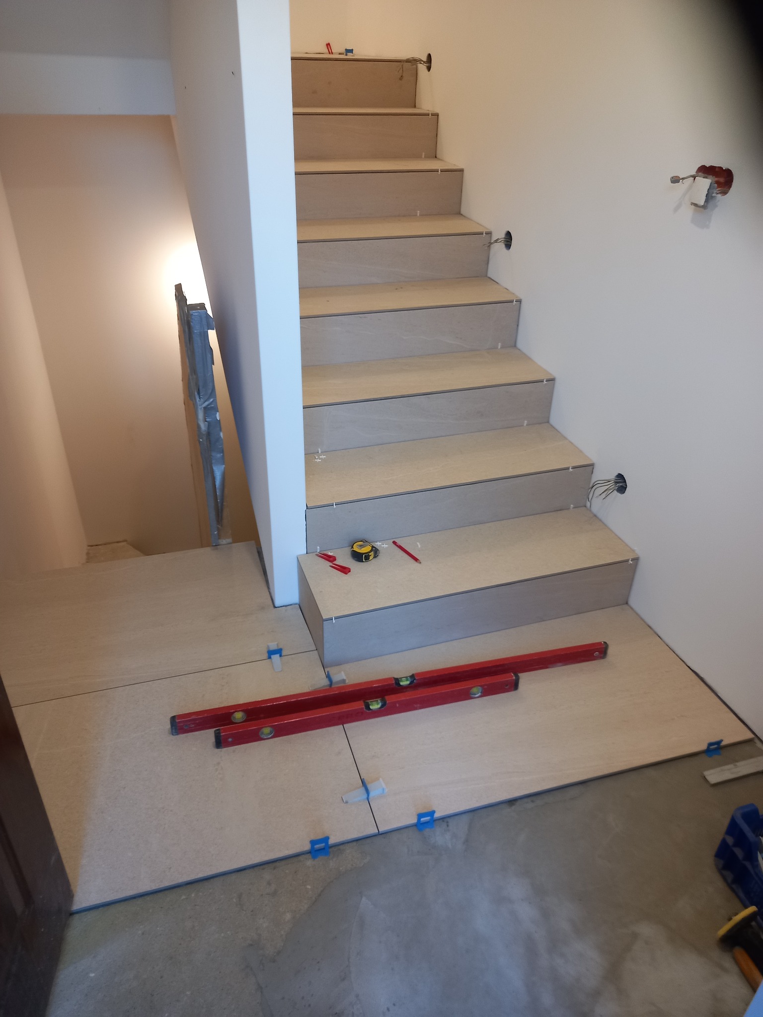 Układanie płytek na schodach z użyciem poziomicy i krzyżyków dystansowych. Widoczne narzędzia glazurnicze i częściowo ułożona podłoga z płytek w neutralnym beżu.