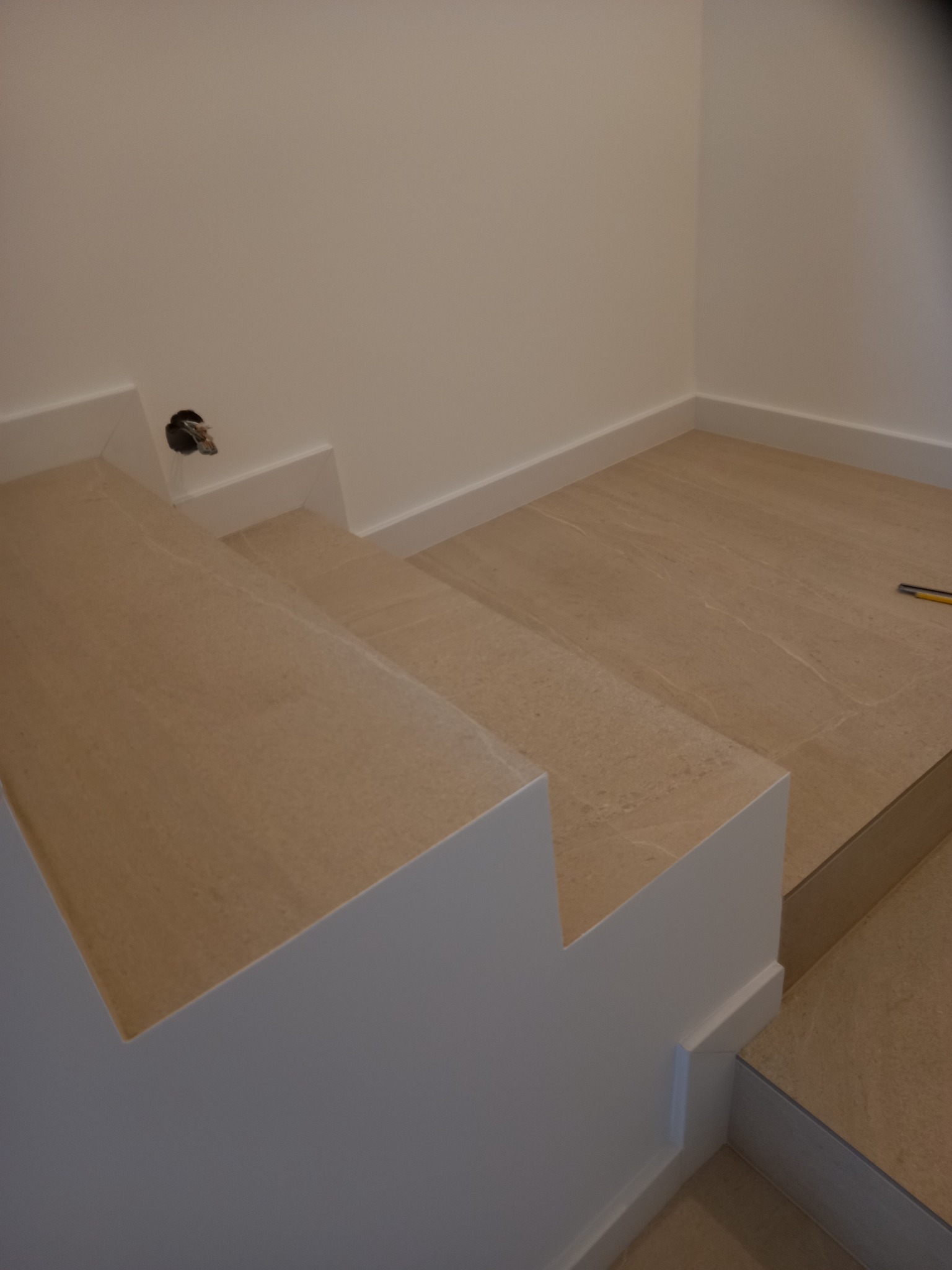 Wykończone schody z beżowych płytek imitujących kamień, białe listwy przypodłogowe i gładkie, białe ściany. Widoczny otwór elektryczny.