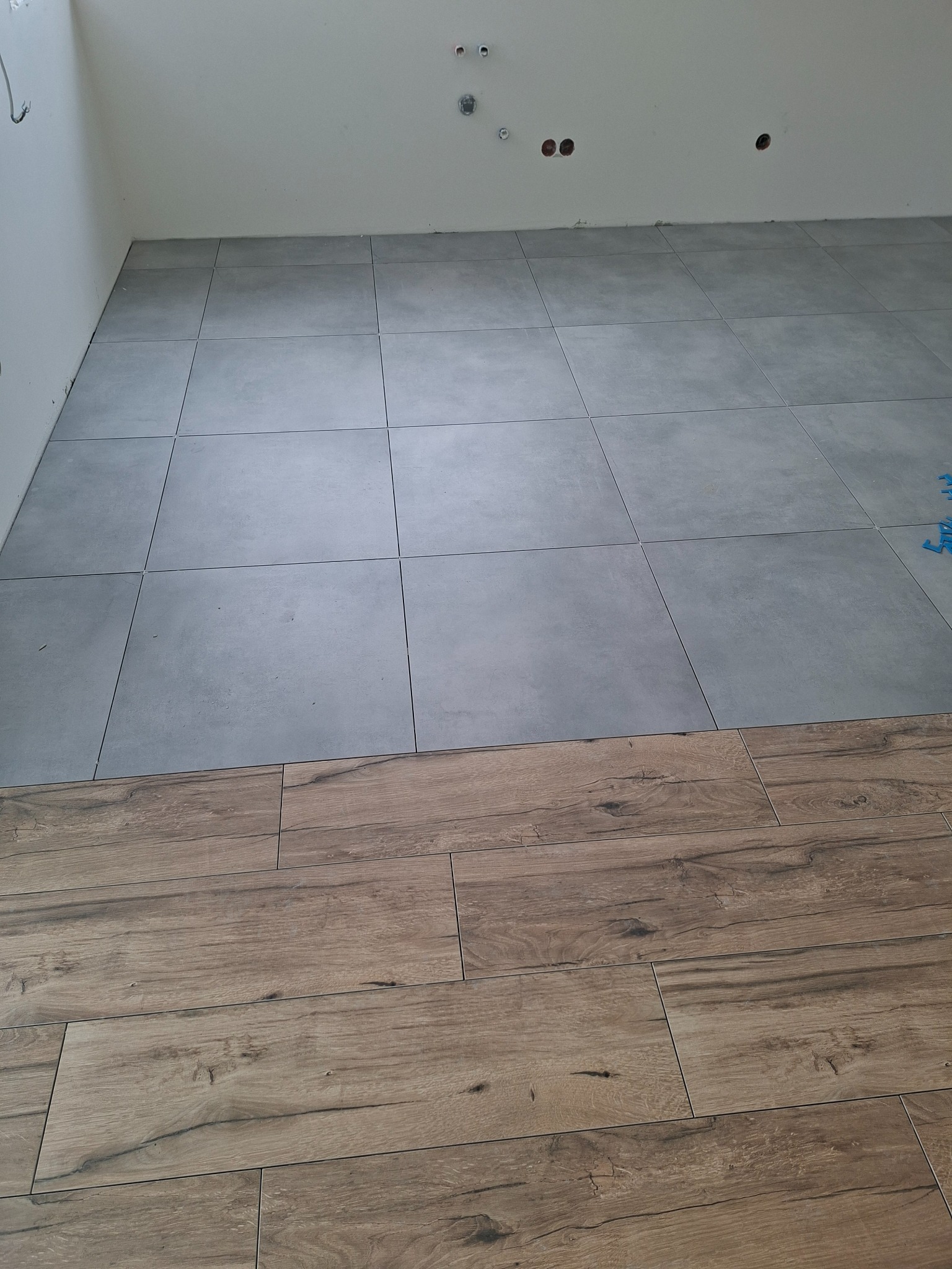 Połączenie szarych płytek podłogowych i paneli imitujących drewno na podłodze w trakcie prac wykończeniowych, widoczne otwory instalacyjne w ścianie.