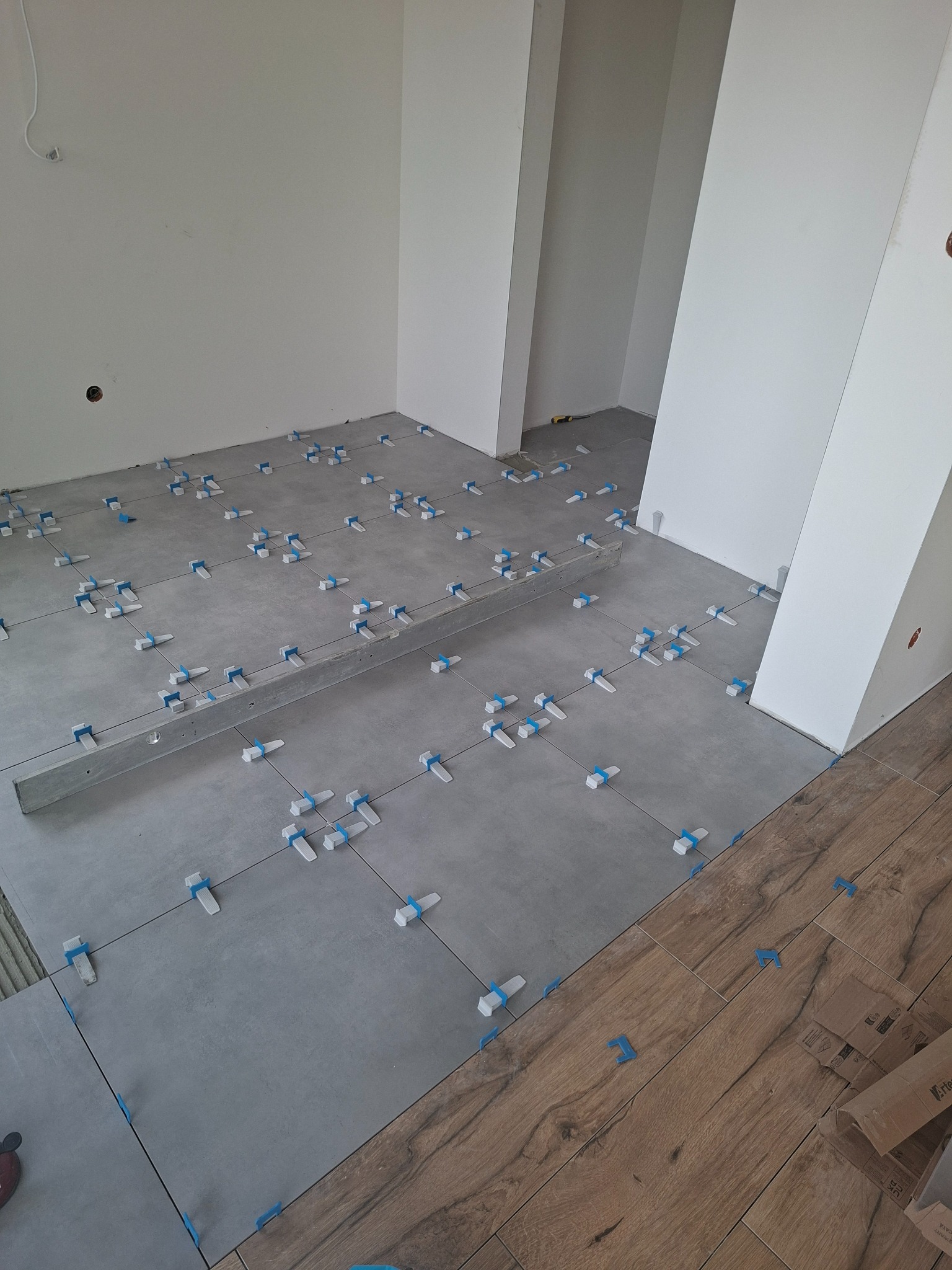 Układanie szarych płytek podłogowych z użyciem poziomicy i systemu poziomowania płytek. Częściowo ułożona drewnopodobna podłoga. Białe ściany w tle.