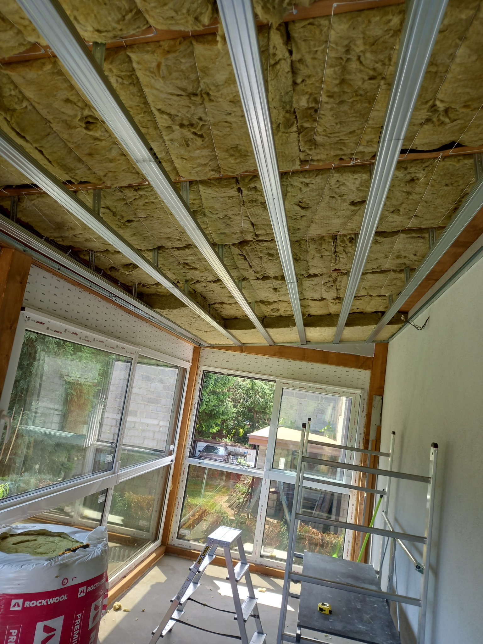 Ocieplenie poddasza wełną mineralną z widoczną konstrukcją metalową. W tle okna z widokiem na zieleń. Na pierwszym planie drabina i materiały izolacyjne Rockwool.