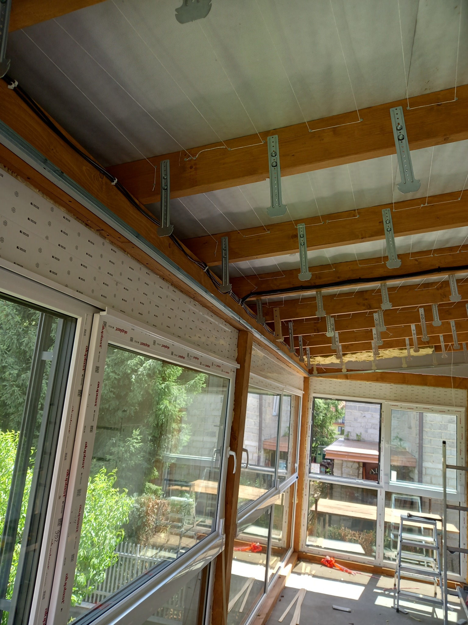 Wnętrze w trakcie prac: drewniana konstrukcja stropu z metalowymi mocowaniami, okna PCV z widokiem na zieleń. Etap przygotowania do ocieplenia poddasza, widoczna drabina.
