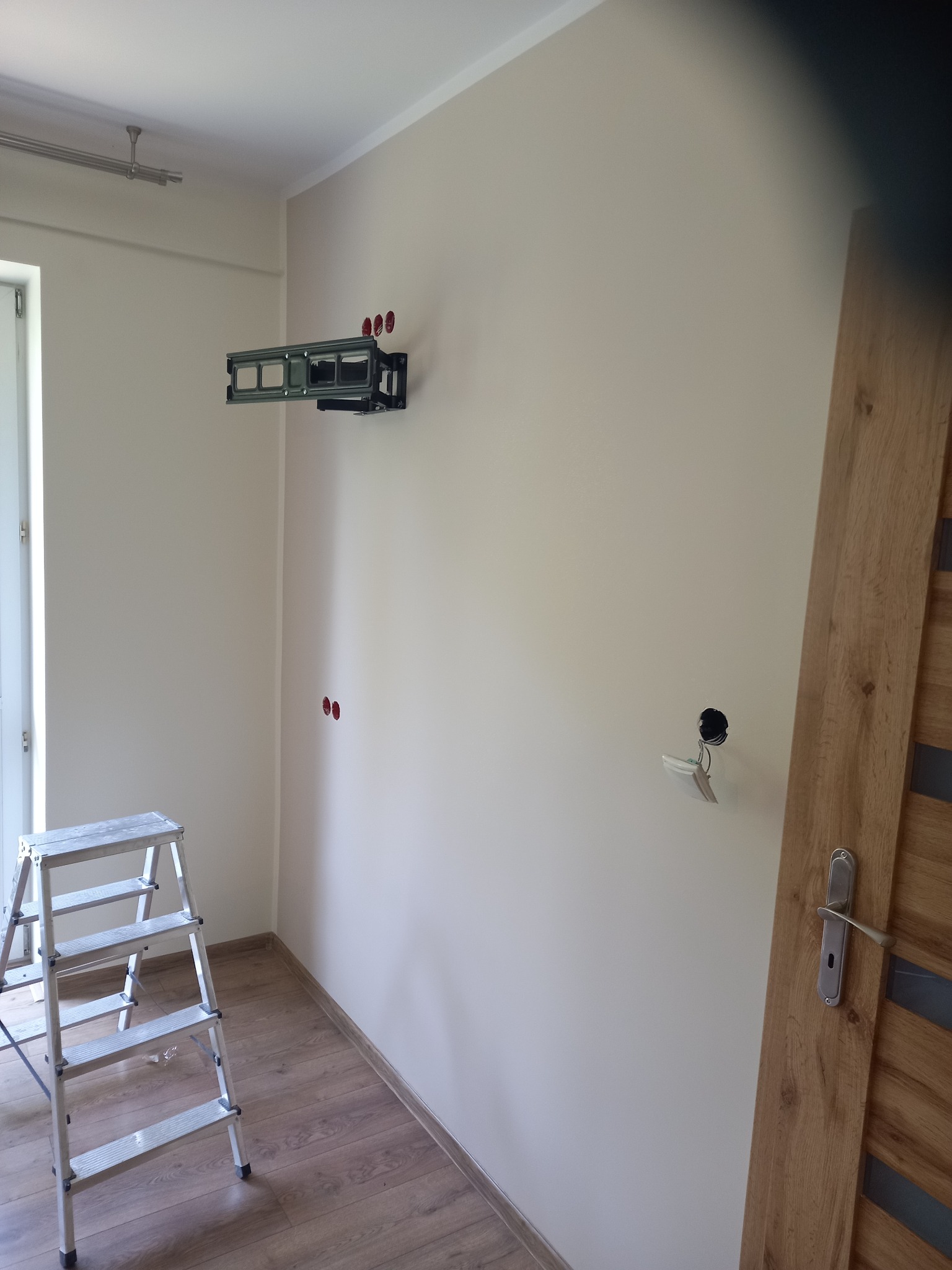 Ściana po malowaniu z zamontowanym uchwytem TV, widoczna drabina malarska, gniazdka elektryczne i fragment drzwi. Parkiet na podłodze.