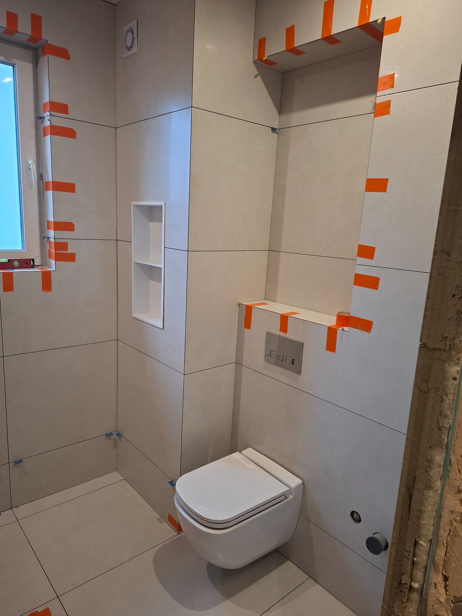 Łazienka w trakcie wykańczania: jasne płytki, zabudowa WC, wnęka w ścianie, okno z poziomicą, zabezpieczenia taśmą. Widoczne detale montażu.