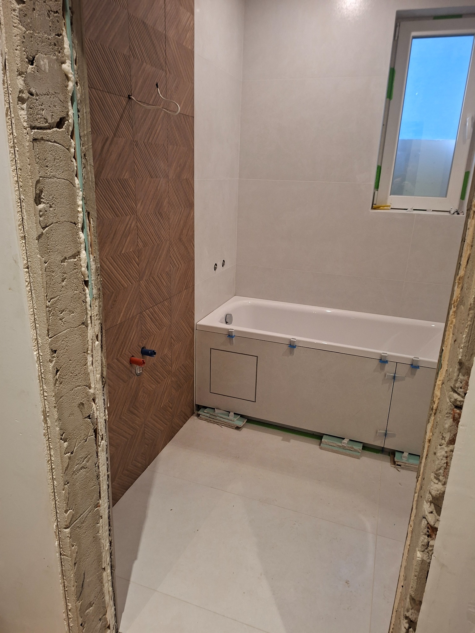 Wnętrze łazienki w trakcie prac wykończeniowych: częściowo ułożone płytki imitujące drewno, wanna obudowana płytkami, okno. Widoczne elementy instalacji i niezakończone obramowanie drzwi.