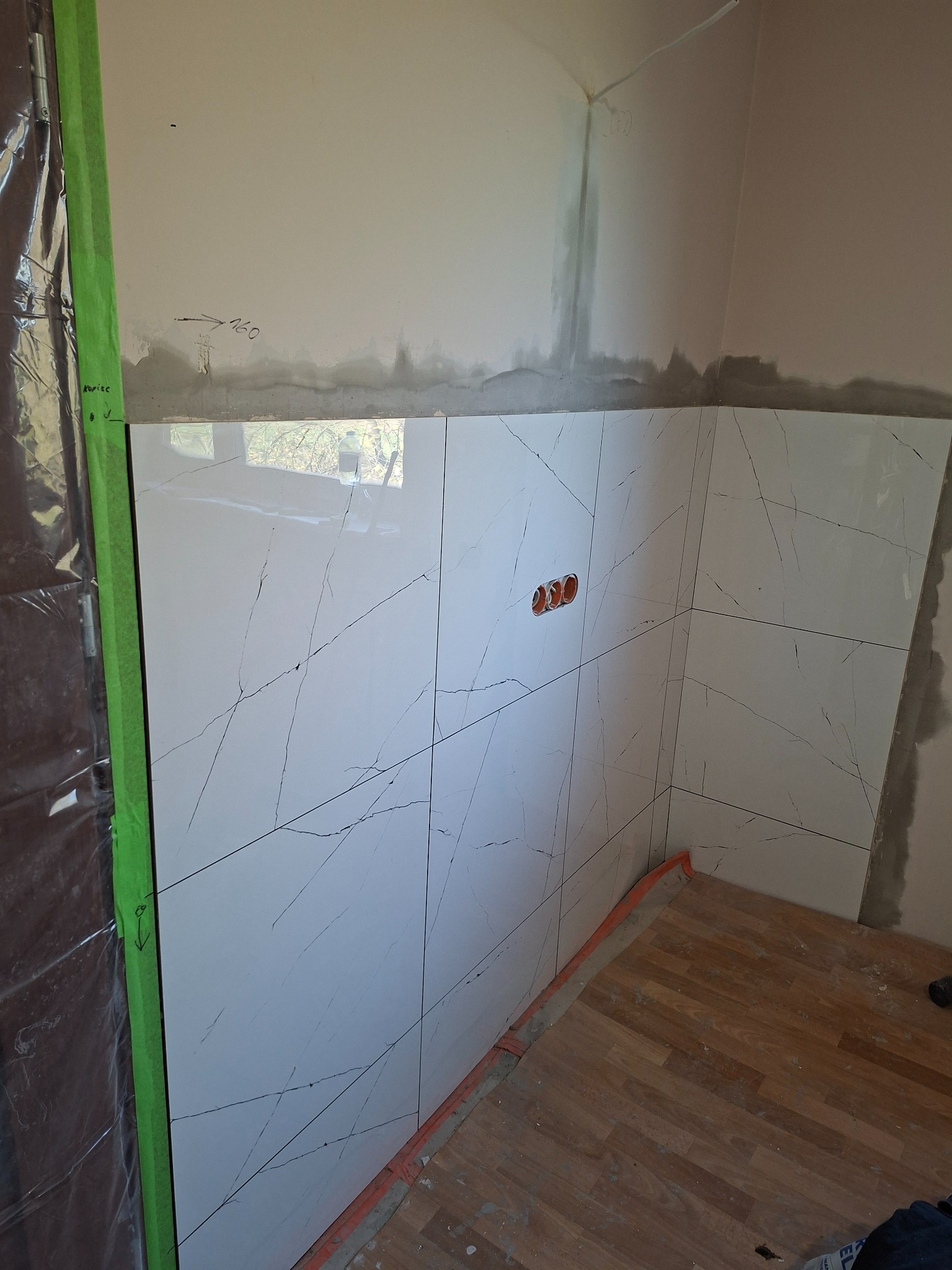 Białe płytki imitujące marmur na ścianie w trakcie montażu, widoczne puszki elektryczne i fragment podłogi z paneli. Prace glazurnicze w trakcie wykańczania.