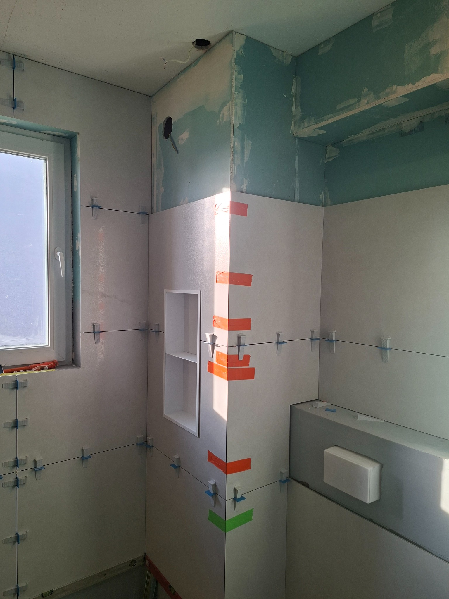 Jasne płytki ścienne w łazience, z widocznymi krzyżykami dystansowymi i taśmą malarską, wnęka w ścianie, okno z widokiem na zewnątrz, surowy stan wykończenia.