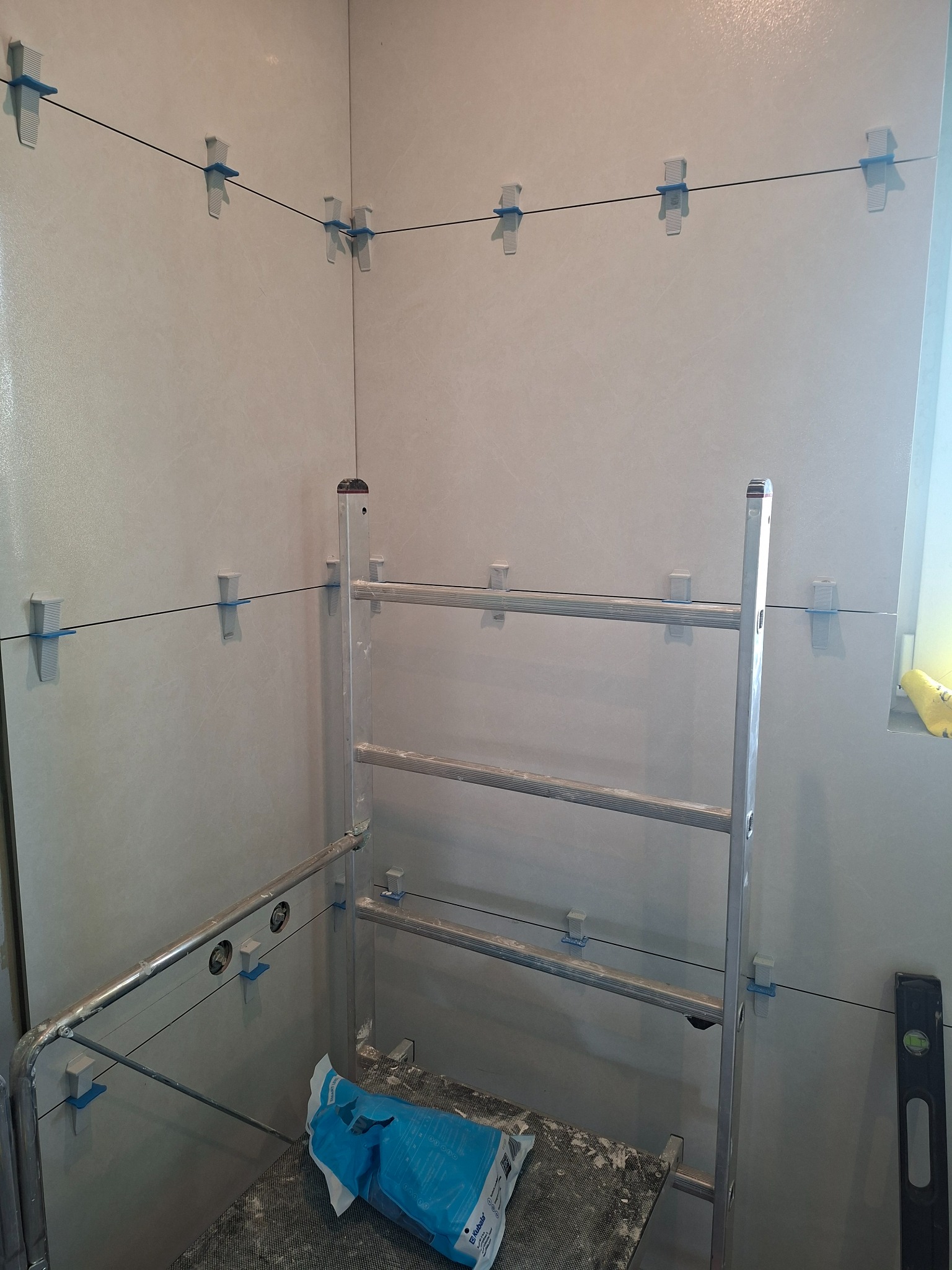 Ściana w trakcie układania płytek, widoczne krzyżyki dystansowe i poziomica. Na drabinie worek z klejem. Praca glazurnicza w toku, jasne płytki.