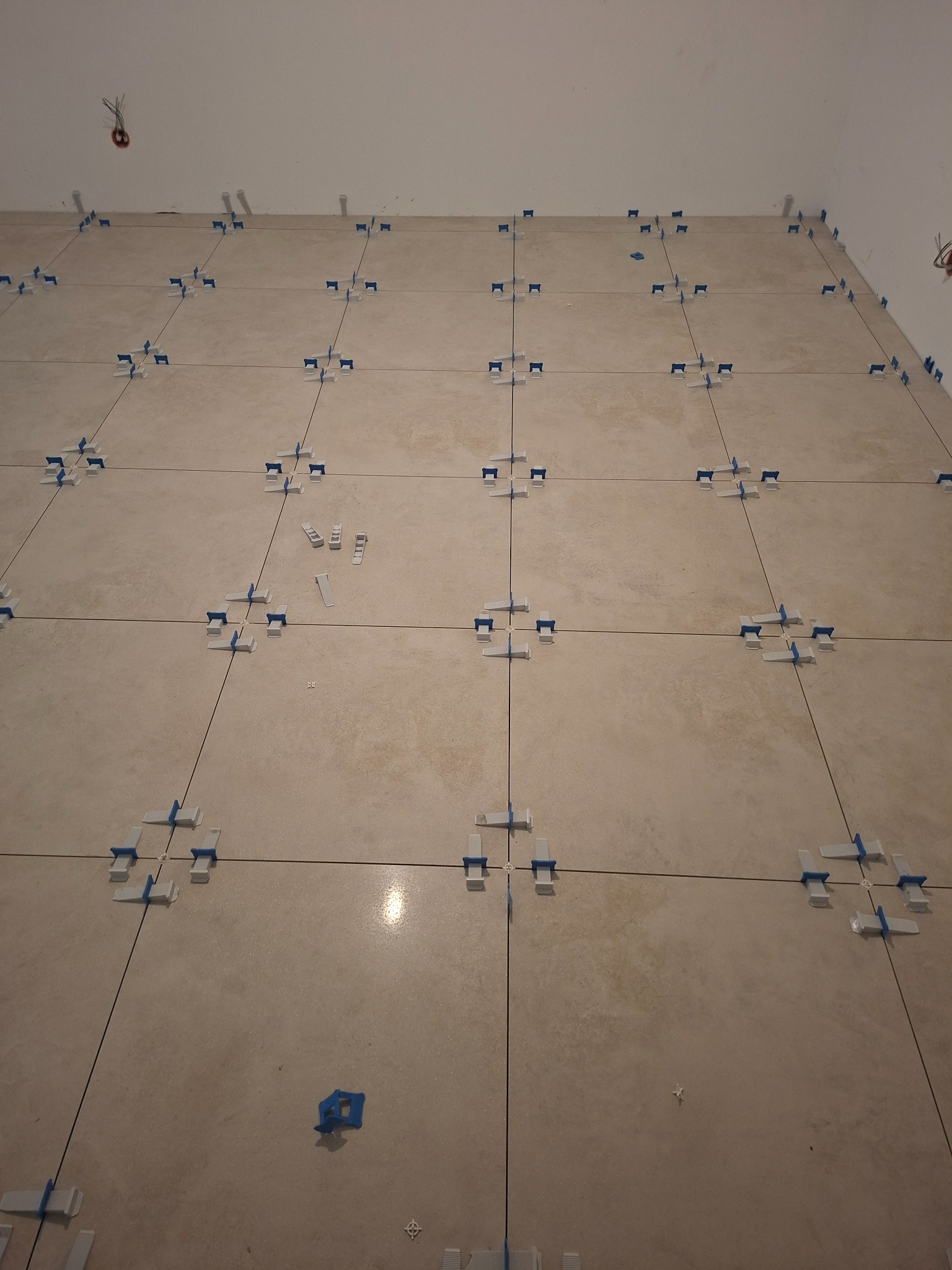 Ułożone beżowe płytki podłogowe z systemem poziomowania, widoczne fugi i elementy systemu. Fragment ściany i wystające przewody elektryczne w tle.