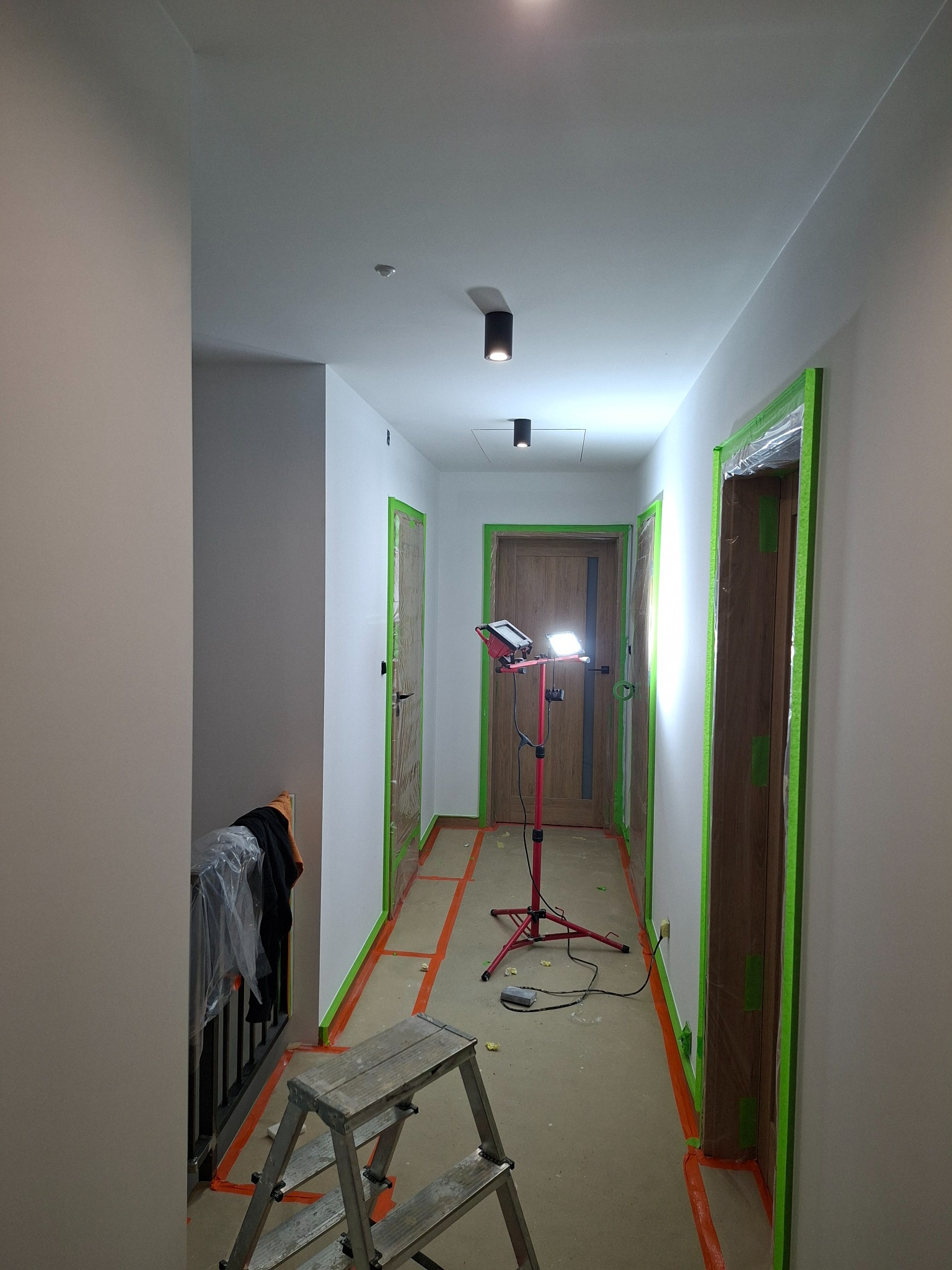Korytarz w trakcie prac przygotowawczych do malowania: ściany zabezpieczone taśmą, drabina, lampa oświetleniowa. Widoczne drzwi z drewnianymi ościeżnicami.